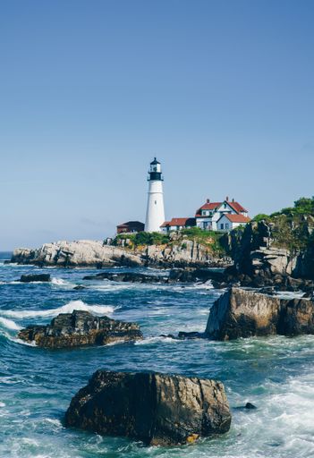 Scenic view of Portland Head Light lighthouse on a rocky coast with waves crashing under a clear blue sky in Cape Elizabeth, Maine