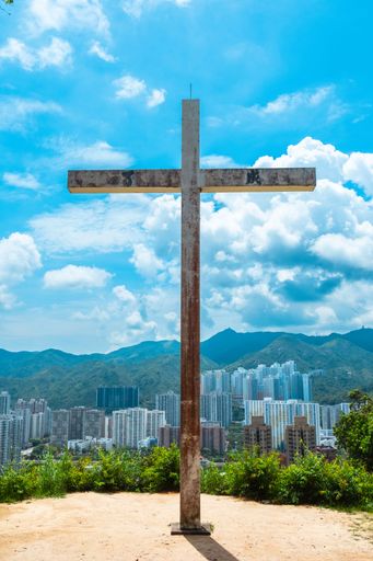A large cross stands against a backdrop of city skyscrapers and mountains under a bright blue sky in Hong Kong.