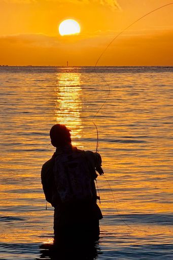 Silhouette of a man fishing at sunset with a vibrant orange sky reflecting on the water.