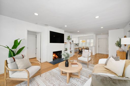 An open-concept living room with a cozy fireplace, wooden flooring, modern furniture, and floor-to-ceiling glass doors leading to an outdoor patio.