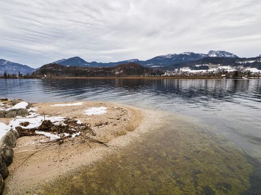 Scenic winter view of a tranquil lake with snow-covered shorelines and distant mountains under a cloudy sky, capturing serene natural beauty.