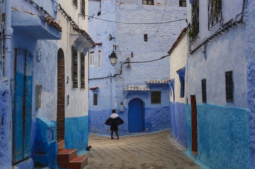 A vibrant street scene in Chefchaouen, Morocco, featuring blue-painted buildings and a person walking, capturing the city's unique charm.