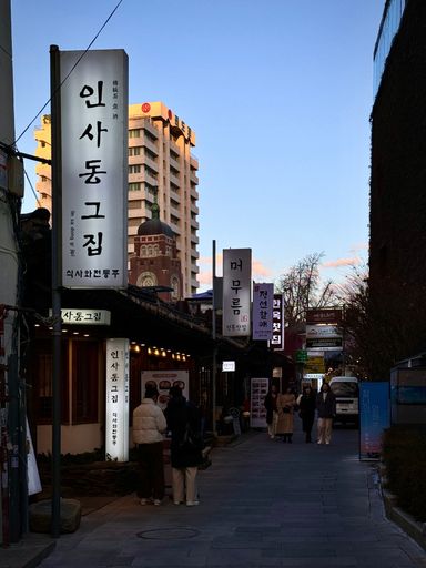 Evening street scene in Insadong, Seoul, with people walking and traditional Korean signs under a clear sky.