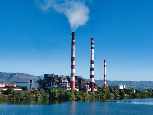 A large industrial power plant with three tall smokestacks emitting smoke into the clear blue sky, situated near a body of water.