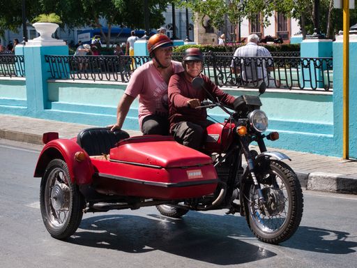 The two men riding a vintage red motorcycle with a sidecar through a sunny city street, evoking nostalgia and classic automotive charm.