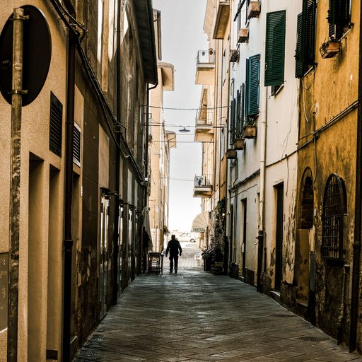 A narrow European alleyway with a lone figure walking, flanked by rustic buildings and balconies on a sunny day.