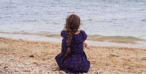 A baby girl with her hair braided wearing a dress sitting on the beach and watching the sea