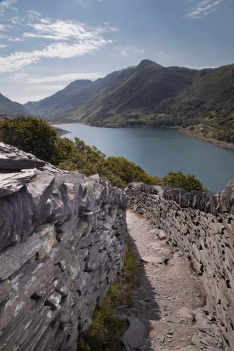 Scenic view of a stone path leading to a tranquil lake surrounded by lush mountains under a clear sky in Snowdonia, Wales.