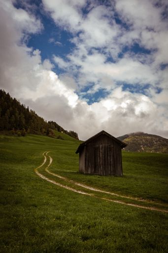 An old wooden barn on a hill covered in greenery under the sunlight and a cloudy sky at daytime
