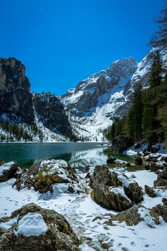 A vertical picture of a lake surrounded by rocks covered in the snow and trees under the sunlight