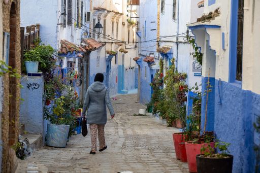 A woman walks through the vibrant blue streets of Chefchaouen, Morocco, surrounded by colorful potted plants and charming architecture