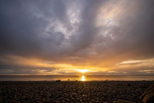 Sunset over a rocky beach with dramatic clouds and golden light reflecting on the water.