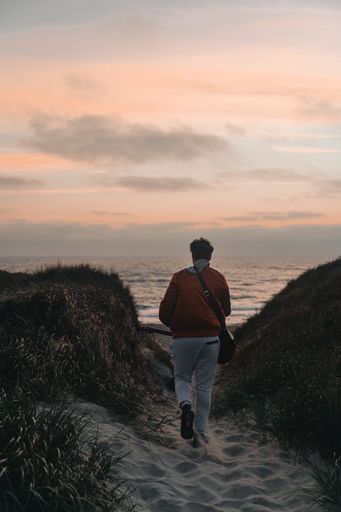 A vertical shot of a male from the back walking on the rocky beach under a cloudy sky at sunset