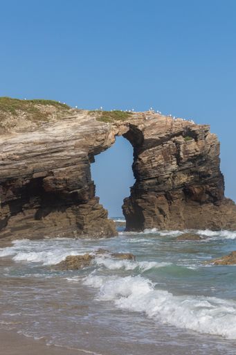 A vertical shot of rocky cliffs sticking out of the sea under a clear sky in As Catedrais, Spain