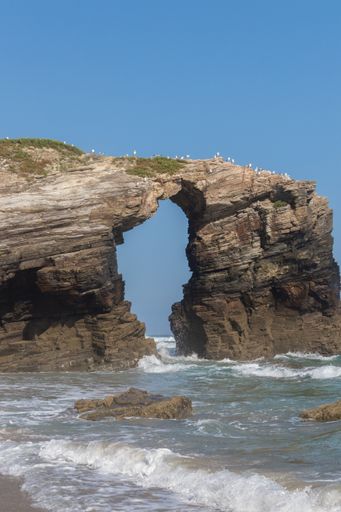 A vertical shot of cliffs sticking out of the sea under a clear sky in As Catedrais, Spain