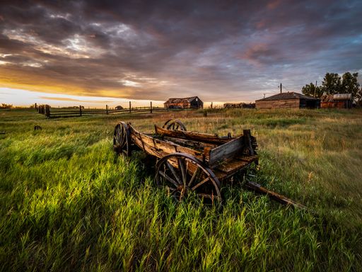 An abandoned wooden wagon on a grassy field under a dramatic sunset sky, with rustic barns in the background, capturing rural tranquility.