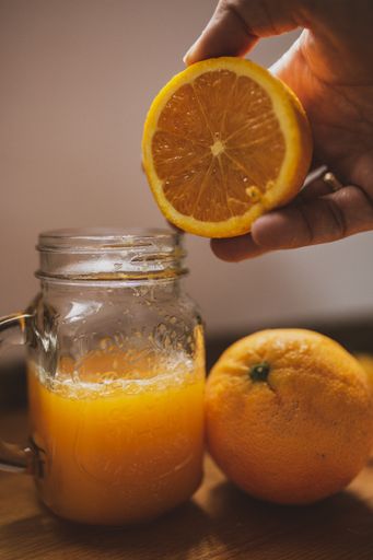 A vertical shot of a person holding an orange near a glass jar filled with orange juice