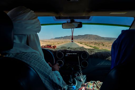 View from inside a car driving through a desert landscape with two people in traditional attire, clear blue sky and distant mountains.