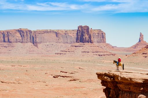 A wide-angle shot of a cool cowboy wearing red shirt, jeans and a white hat sitting on a horse on the edge of a cliff in Monument Valley