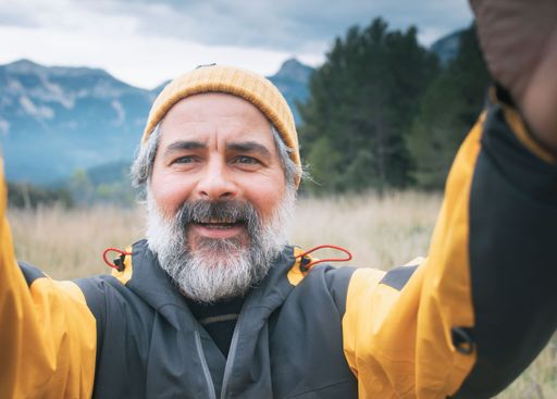 Middle-aged man with a beard taking a selfie in a mountainous landscape wearing a yellow jacket and beanie.