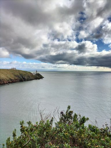 Scenic view of a coastal landscape with a lighthouse under a dramatic cloudy sky, featuring lush greenery in the foreground.
