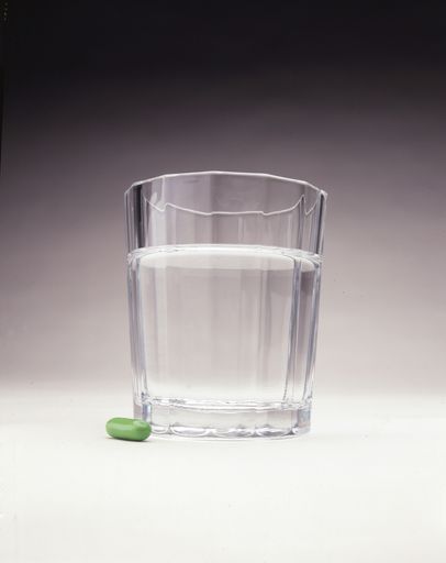 A vertical shot of a green pill near a glass of water on a white surface
