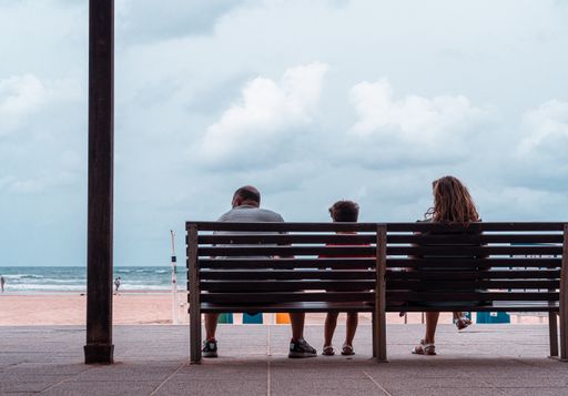 A family sitting on a wooden bench and looking at the sandy beach captured from behind