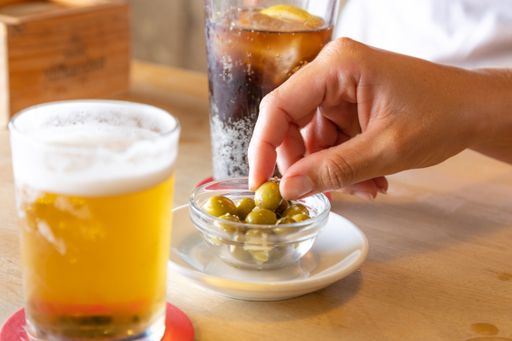 A human hand taking green olive in glass bowl between two cups of drinks on wooden table