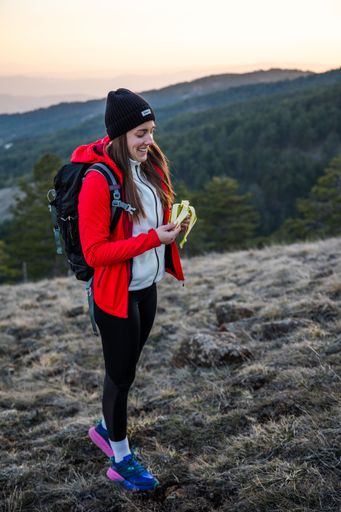 Young woman in hiking gear enjoys a snack on a mountain trail at sunset, wearing a red jacket and beanie, with a scenic forest view.