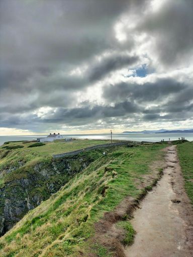 Scenic coastal landscape with a dramatic cloudy sky over a cliffside path leading to a distant house, capturing the natural beauty and tranquility.