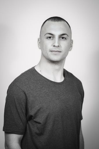 A grayscale portrait of a handsome multi-racial man with a buzz cut standing against the wall