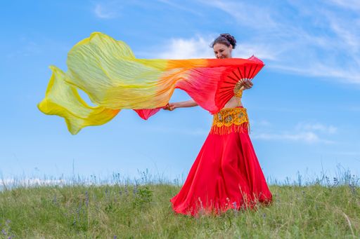 Young woman in colorful dress is dancing fascinating oriental dance with bright yellow-red fans on green meadow against blue summer sky.