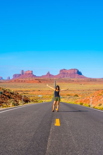 A vertical shot of a young female standing in the middle of the road with open arms in Monument Valley, USA