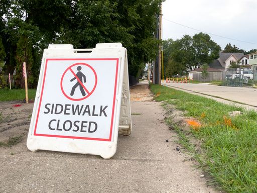 A sidewalk closed sign by the road and lawn on the background of houses and trees