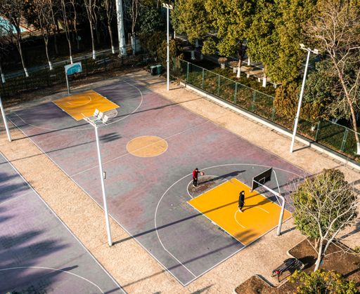 Aerial view of an outdoor basketball court with two people playing, surrounded by trees and pathways in a park setting.