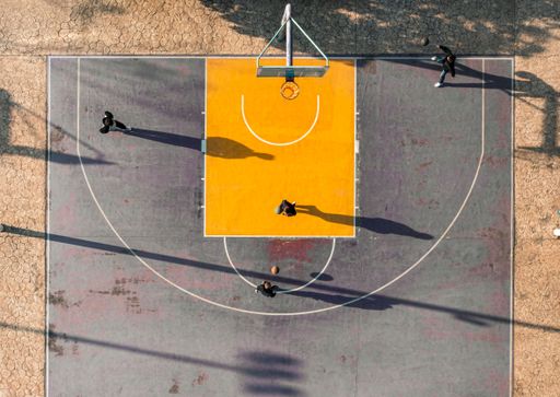 Aerial view of a basketball court with players casting long shadows during a game on a sunny day.