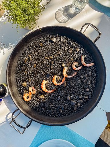 Traditional black seafood paella with shrimp and squid ink, served in a pan on a sunlit table with a fresh and Mediterranean setting.