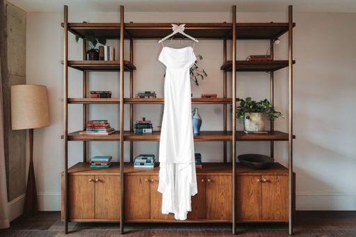 A white wedding dress hanging on a wooden shelf with books and plants, creating a cozy interior setting.
