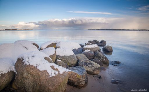 Snow-covered rocks by a tranquil sea with distant islands under a partly cloudy sky.