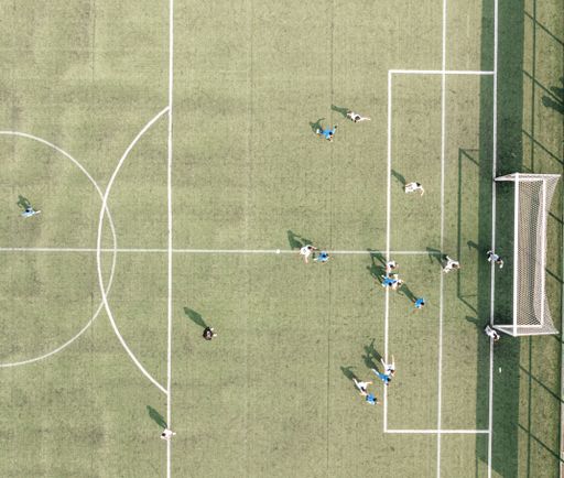 Aerial view of a soccer match on a green field with players in action, casting long shadows in the sunlight.