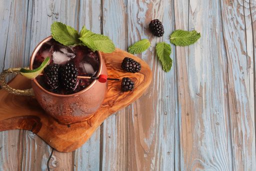 A metal cup of a refreshing drink with mint, lime and blackberries on a wooden coaster