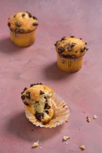 Three chocolate chip muffins on a textured surface, with one partially eaten, showcasing a delicious baked treat.