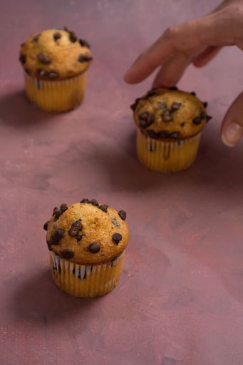 Three chocolate chip muffins on a textured surface with a hand reaching for one, creating a cozy and inviting atmosphere.