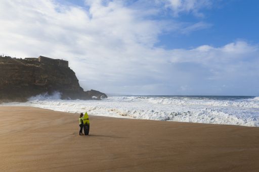 A surfer standing on a sandy shore and looking at a waves crashing on the ocean with a coastal cliff in the background