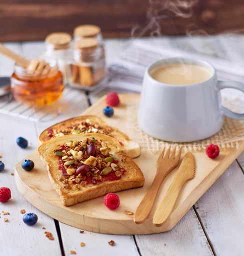 A closeup of toasts with jam and nuts on a wooden board with a cup of hot coffee on it