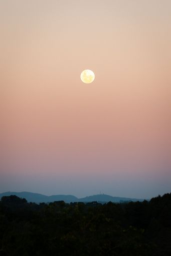 The Full moon rising over a serene landscape with a pastel sky and distant hills at dusk.