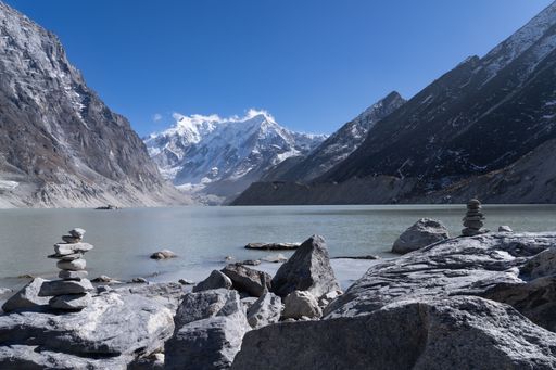 Scenic view of a tranquil mountain lake with stone cairns and snow-capped peaks under a clear blue sky.