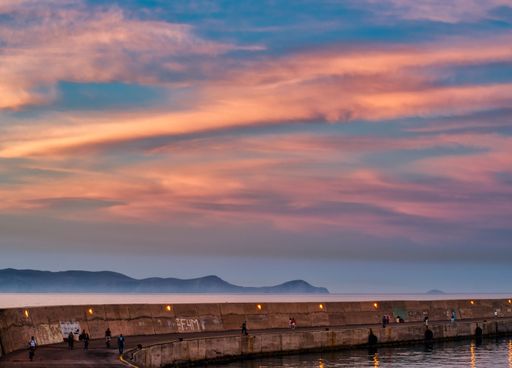 A scenic view of a pier at sunset with colorful clouds and distant mountains, people walking and enjoying the serene atmosphere.