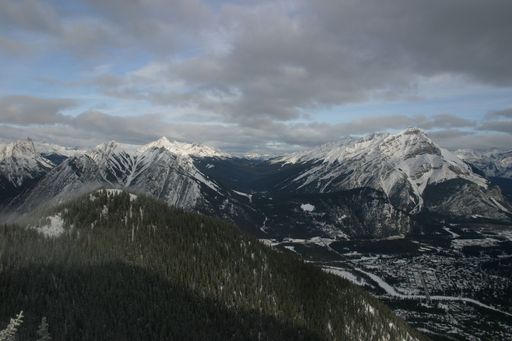 A panoramic view of snow-capped mountains and a forested valley under a cloudy sky in Banff National Park, Canada.