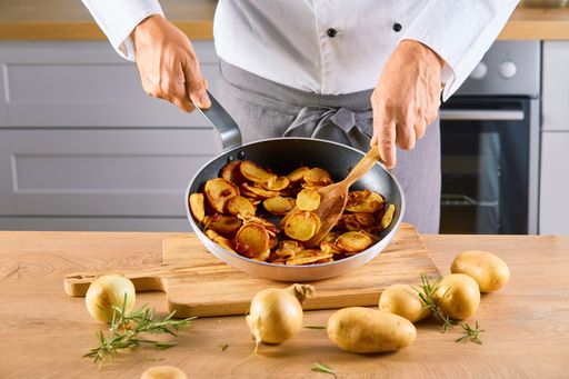 A male cook holding a pan of fried potatoes with meat slices and mixing with a spatula on a board